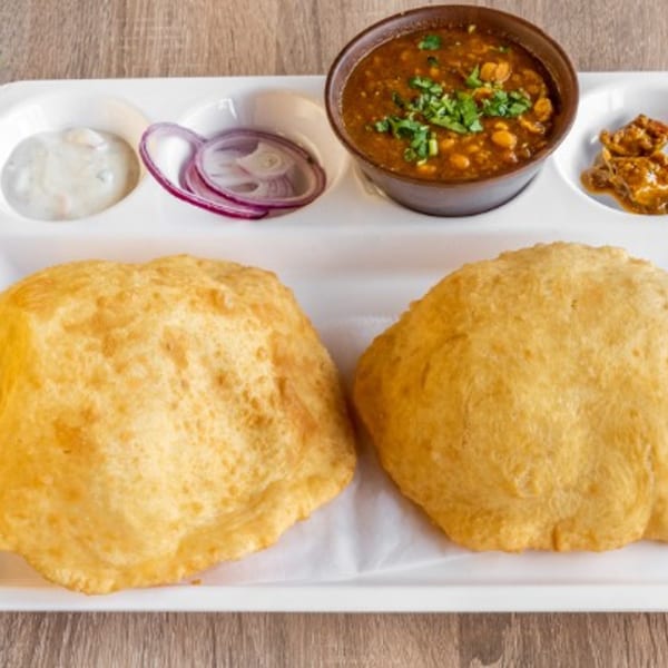 Two golden-brown Indian puri breads with chana masala, yogurt, red onions, and pickle on a white tray.