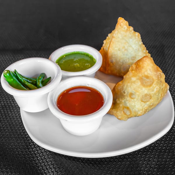 Two golden-brown samosas with three small white bowls of green chutney, red sauce, and green chilies.