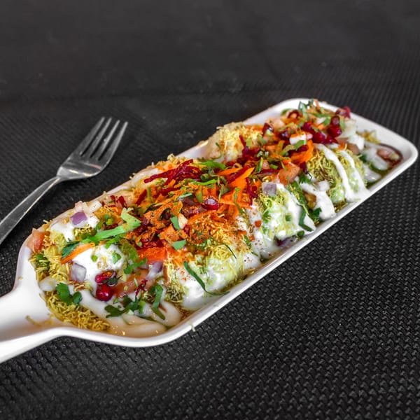 A long white plate of Indian chaat, topped with colorful vegetables, sauces, and spices, with a fork.